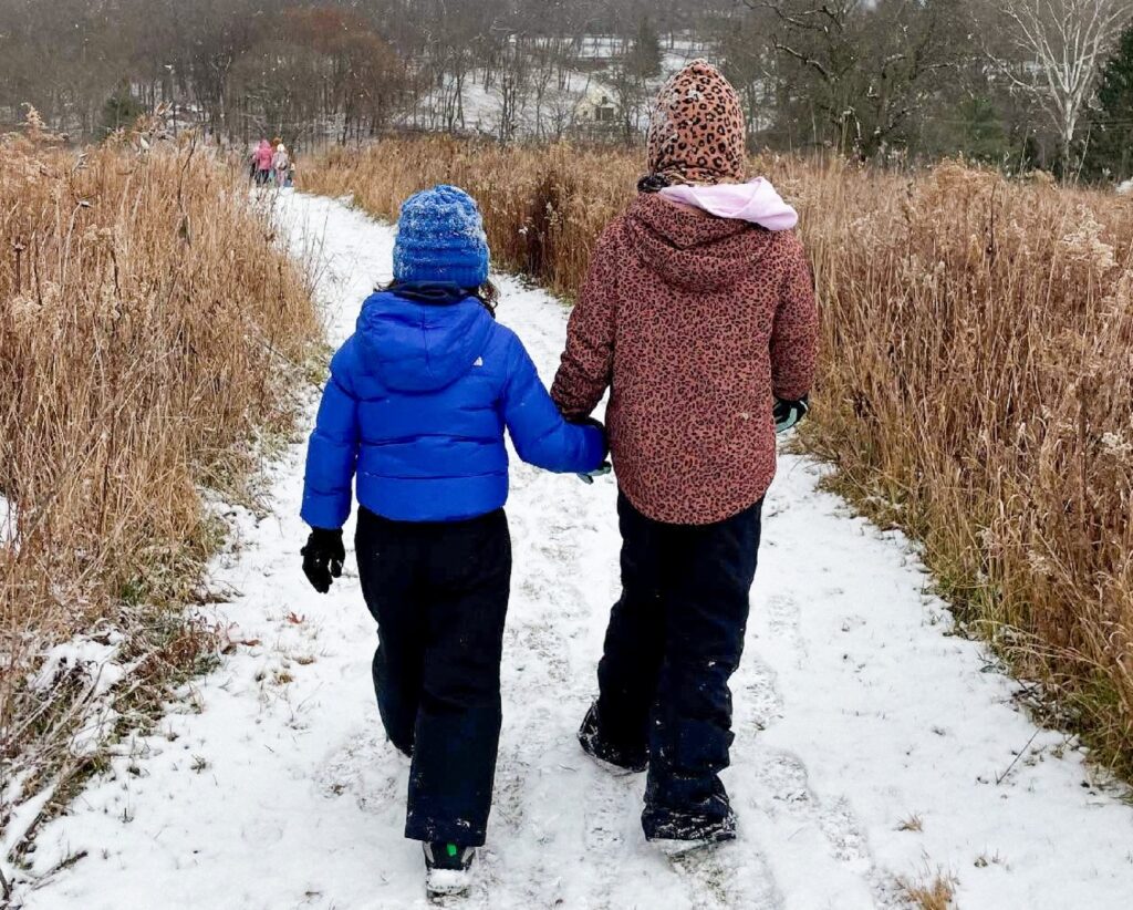 Two children walk hand in hand along a snowy path between tall dried grasses, in blue and leopard-print coats.