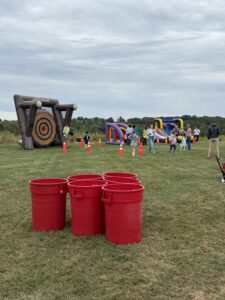 Group of people at an outdoor fair with inflatable obstacle courses, orange cones, and large red plastic bins in a grassy field.