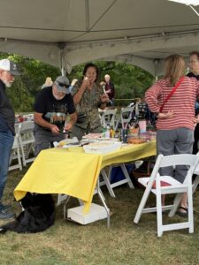 Outdoor gathering under a tent with people around a long table of food; a man in a black t‑shirt adjusts items, a woman tastes something, a large black dog sits at the edge of the frame.