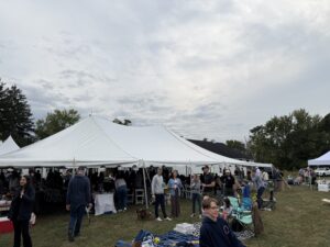 Outdoor event under large white tents with people mingling and tents lining a grassy field under a cloudy sky, some children toys visible in the foreground.