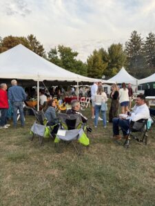 People socialize under white canopy tents at an outdoor event on a grassy field.
