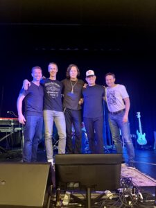 Five men stand arm-in-arm on a stage after a performance, with instruments and gear in the background (keyboard left, guitars right).